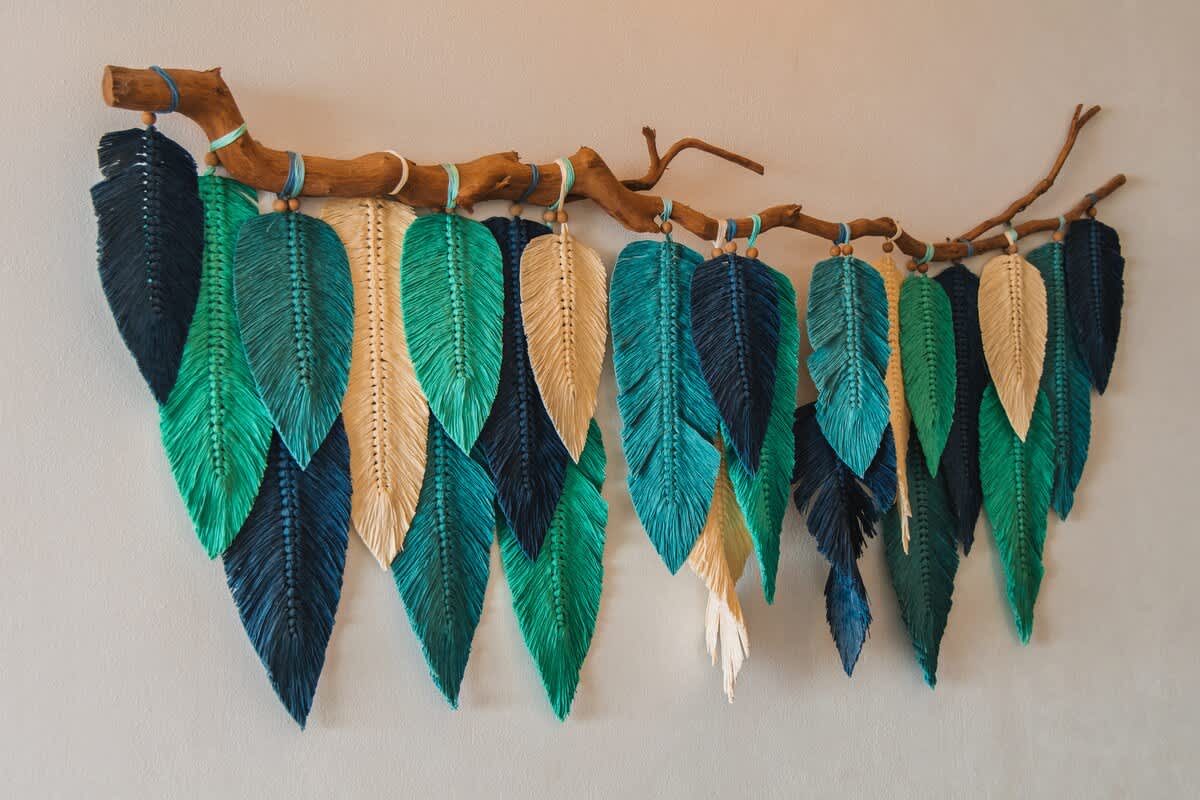A wide shot of a colorful macrame wall hanging, showcasing an array of feather-shaped elements in varying shades of blue, green, and natural beige, suspended from a rustic wooden branch against a light wall, enhancing the room's aesthetic.