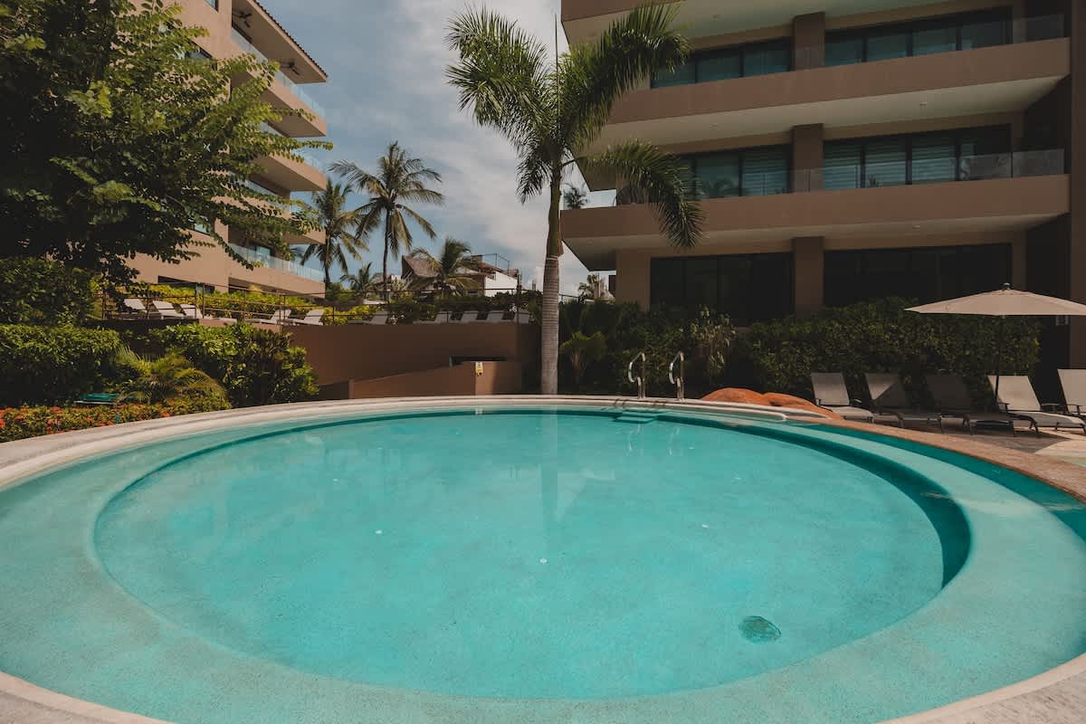 A large, inviting circular swimming pool with clear blue water, surrounded by lounge chairs and lush tropical landscaping. Modern apartment buildings rise in the background under a bright sky.