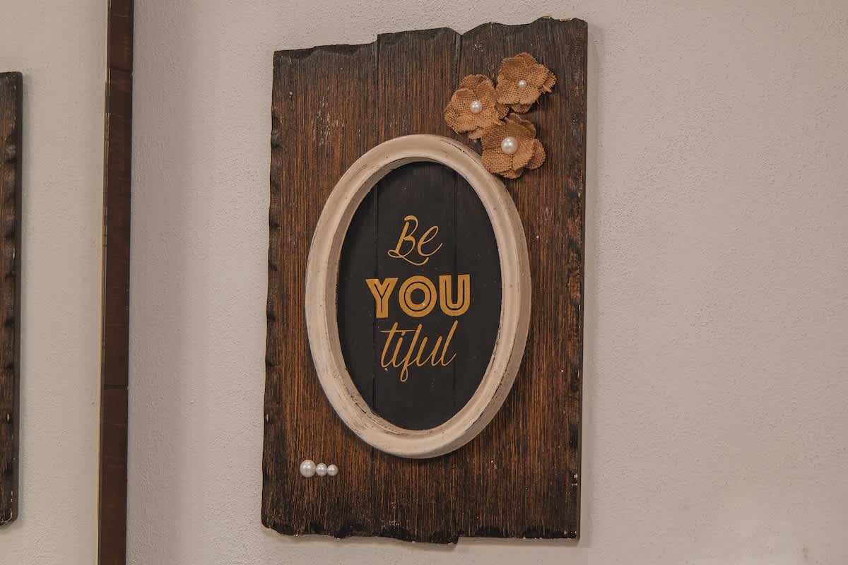 Rustic wall art features a wooden plank background with an oval frame enclosing the word "Be YOU tiful" in gold lettering. Decorative burlap flowers and small pearls adorn the piece.