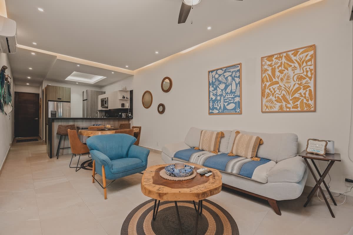 An open-concept living area with a light gray sofa, a blue armchair, and a unique round wooden coffee table on a circular rug. The space flows into a dining area and a modern kitchen with a breakfast bar.