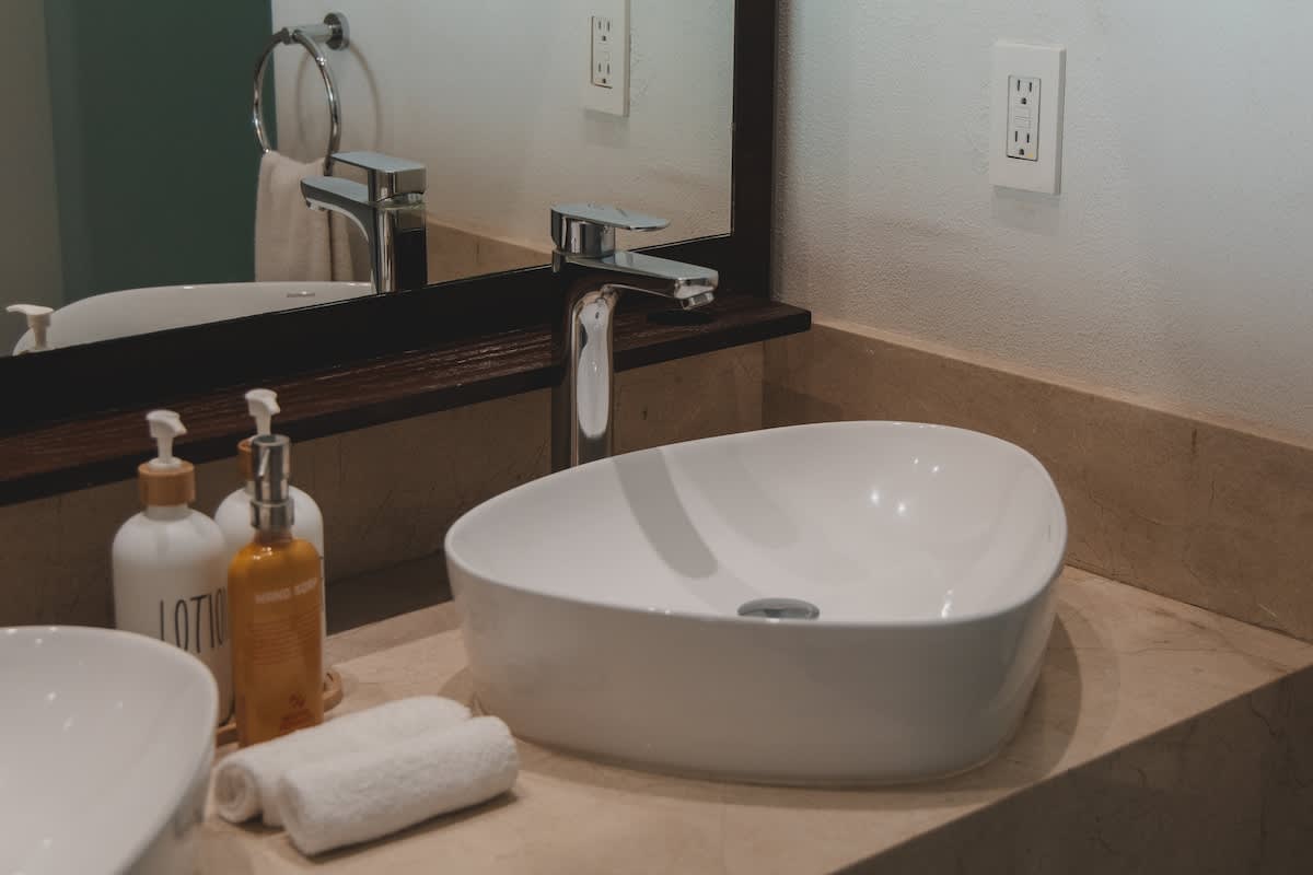 A modern white vessel sink with a chrome faucet sits on a light-colored stone countertop, reflecting in a large mirror, with white towels and soap dispensers visible.
