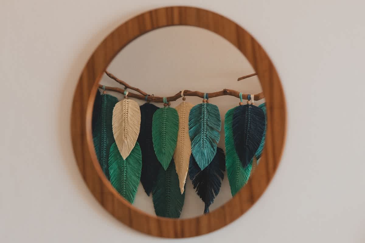 A round wooden-framed mirror reflects a decorative macrame wall hanging featuring various shades of green and beige leaf-shaped elements suspended from a branch, adding a natural, bohemian touch.