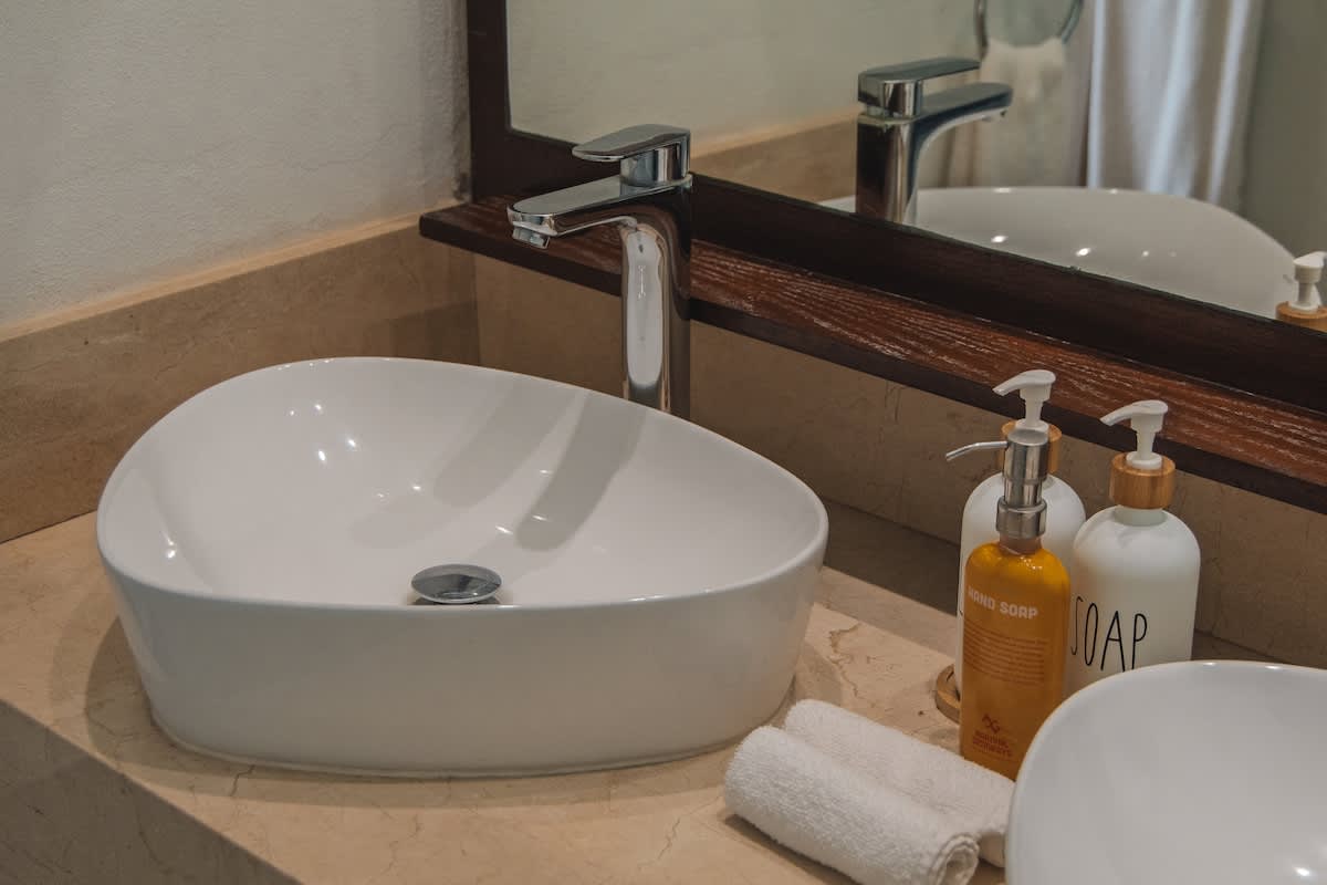 A close-up of a sleek white vessel sink and a tall chrome faucet on a light marble countertop, with various soap and lotion bottles and rolled white towels neatly arranged beside it.