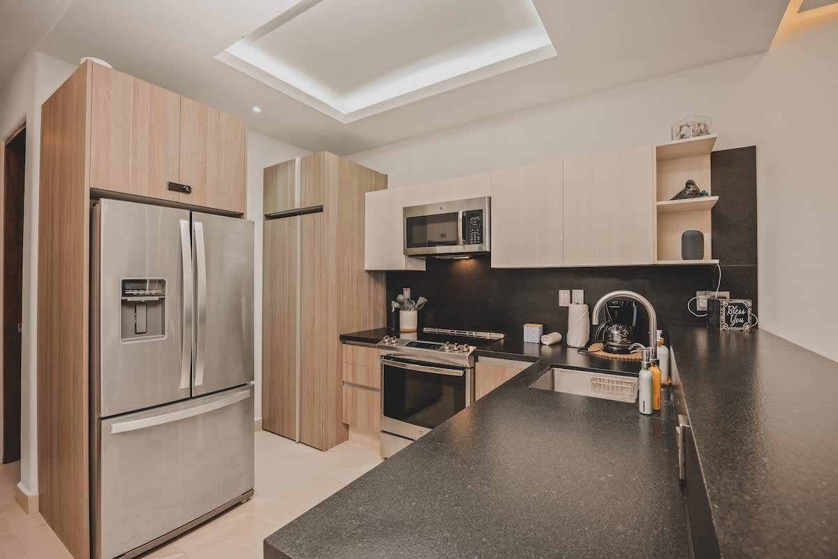 Close-up view of the modern kitchen, highlighting the stainless steel refrigerator, light wood cabinets, dark countertops, and a built-in microwave above the electric range. Various kitchen essentials are neatly arranged.