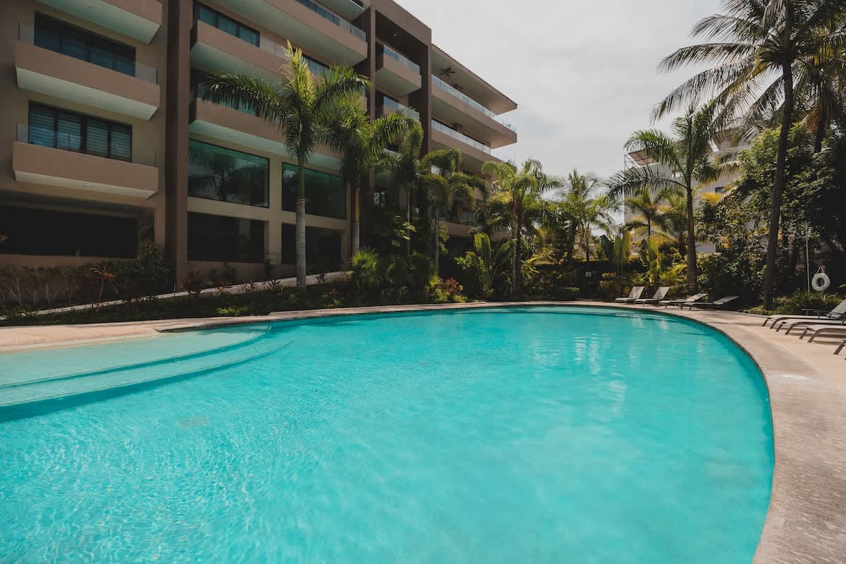 A large, irregularly shaped swimming pool with clear blue water, bordered by concrete decking and lush tropical foliage, with modern apartment buildings and palm trees in the background.
