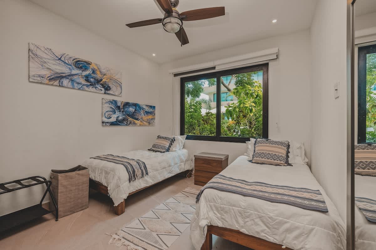 A bright bedroom featuring two twin beds with white bedding and blue patterned throws, a dark wood ceiling fan, and a large window overlooking lush greenery. Abstract blue and gold artwork adorns the white walls.