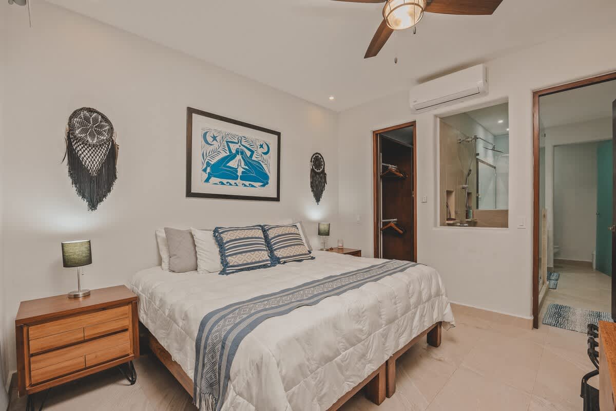 A serene bedroom featuring a king-sized bed with white bedding and a blue patterned throw, flanked by wooden nightstands with lamps. A large framed artwork hangs above the bed, and a ceiling fan is visible.