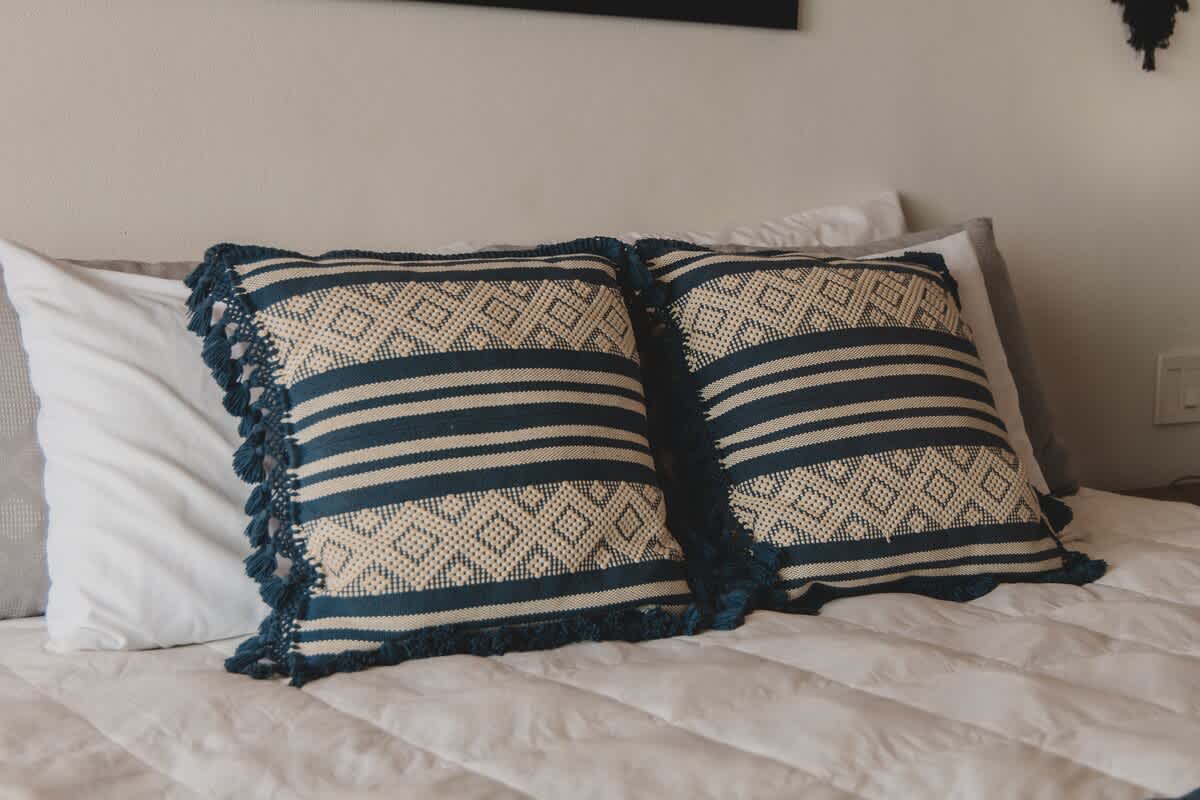 Two patterned pillows with blue and beige designs and fringed edges are neatly arranged on a white quilted bed, with plain white pillows visible behind them.