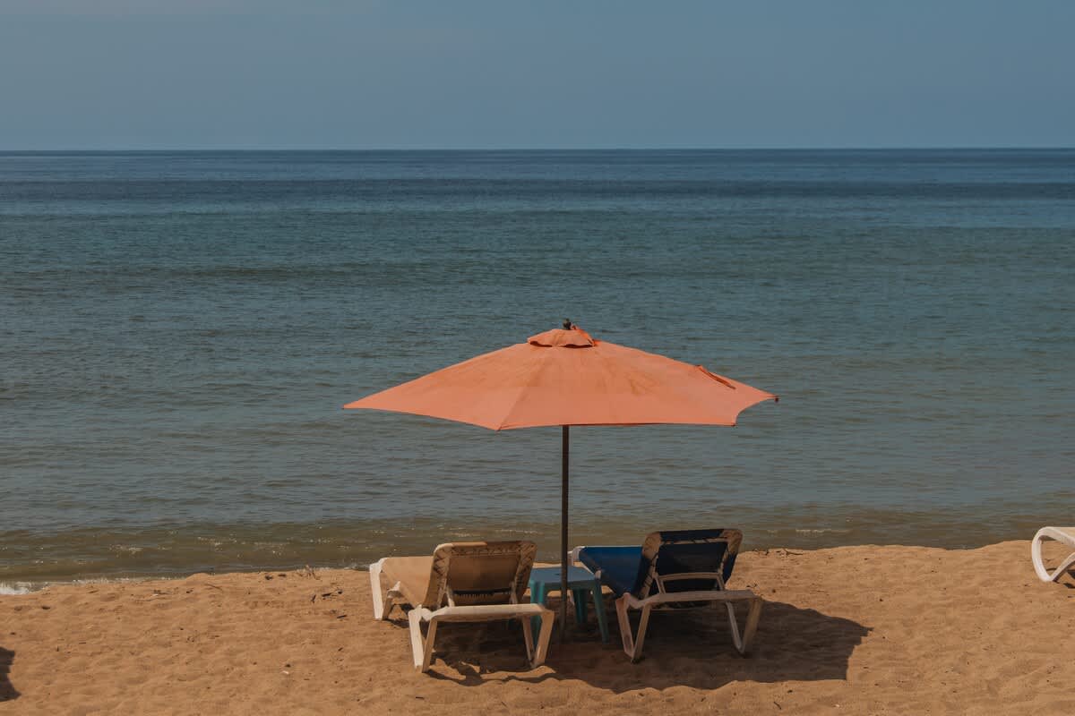 A serene beach scene with two lounge chairs and a large orange umbrella providing shade on the sandy shore, facing the calm blue ocean under a clear sky, highlighting the property's beachside location.