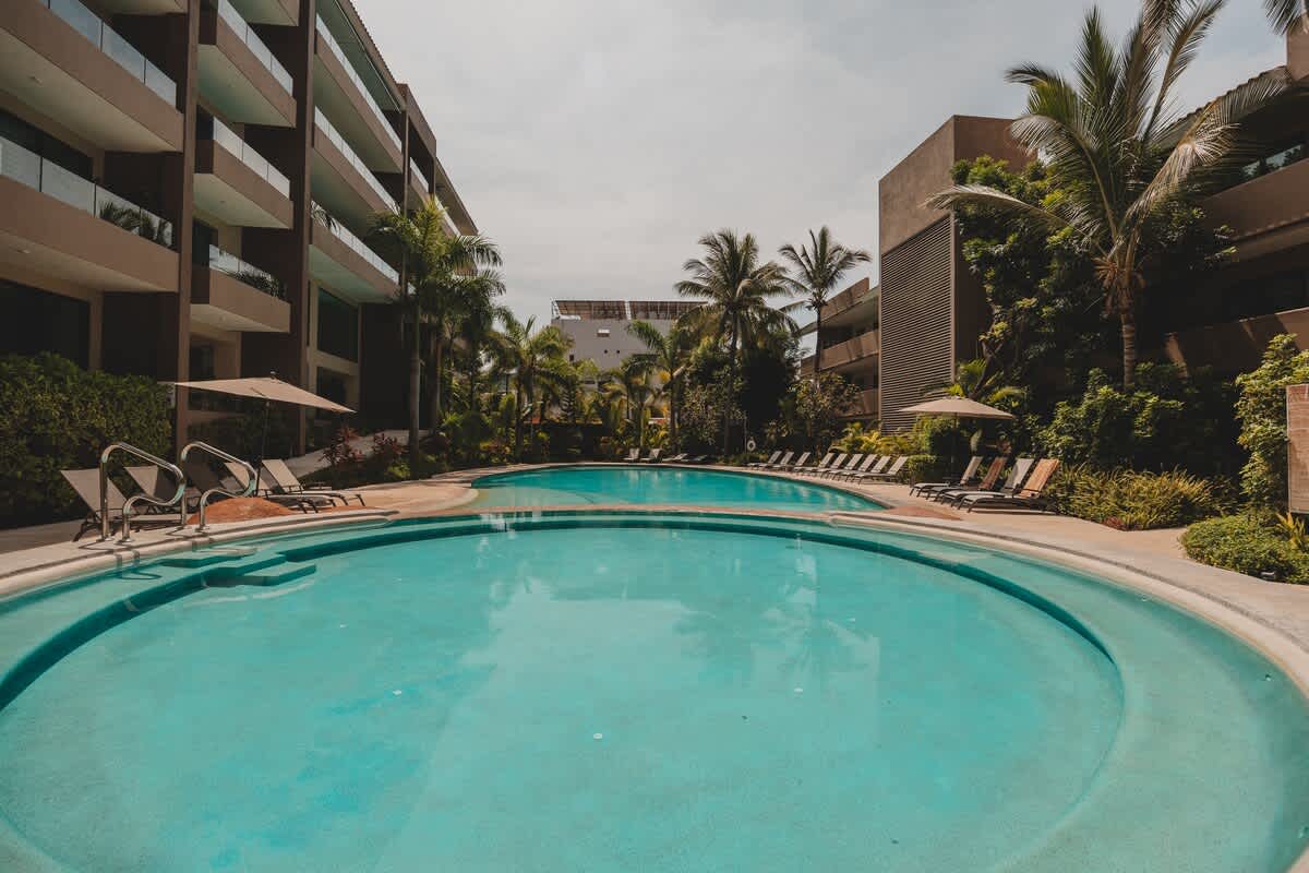 An expansive outdoor pool area featuring multiple interconnected swimming pools with light blue water, surrounded by numerous lounge chairs, tropical plants, and modern multi-story buildings.