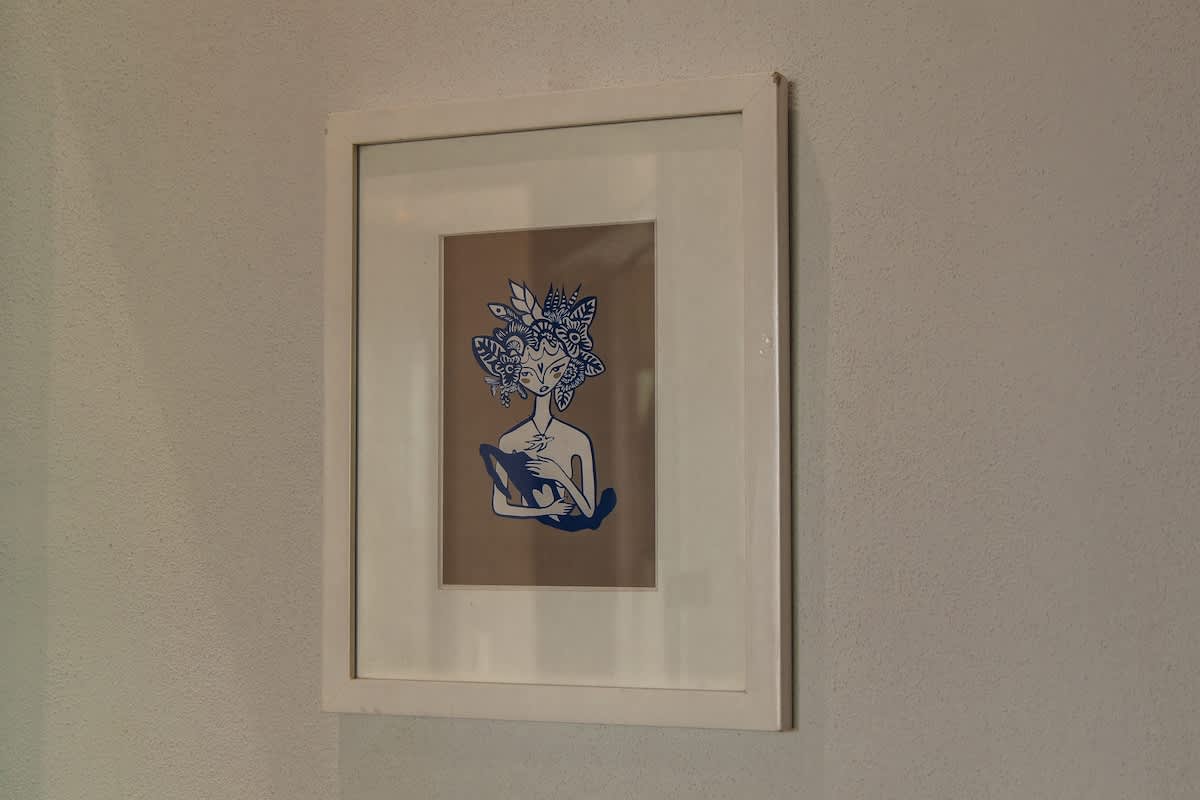 A framed artwork features a stylized figure with an elaborate headdress and holding a small animal, rendered in blue on a beige background, set within a white frame on a textured wall.