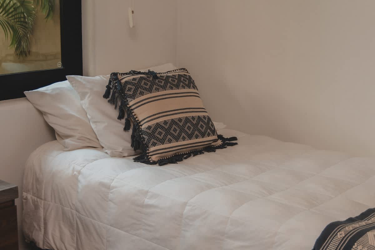 A comfortable single bed dressed in white quilted bedding, featuring a stylish throw pillow with a black and white patterned design and tassels. A dark window frame and a wooden nightstand are visible.