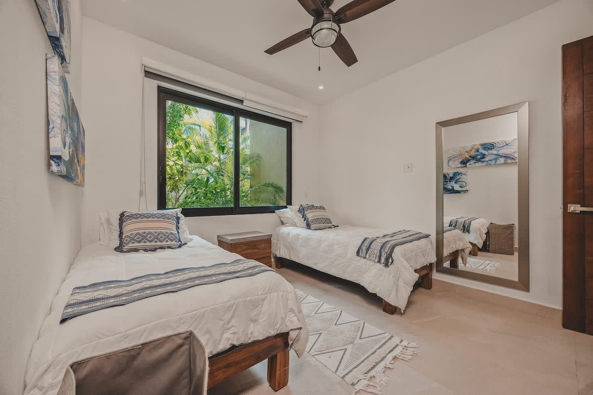 Bright bedroom with two twin beds featuring white bedding and blue patterned throws, a large window overlooking tropical foliage, and a full-length mirror. A dark wood nightstand sits between the beds.