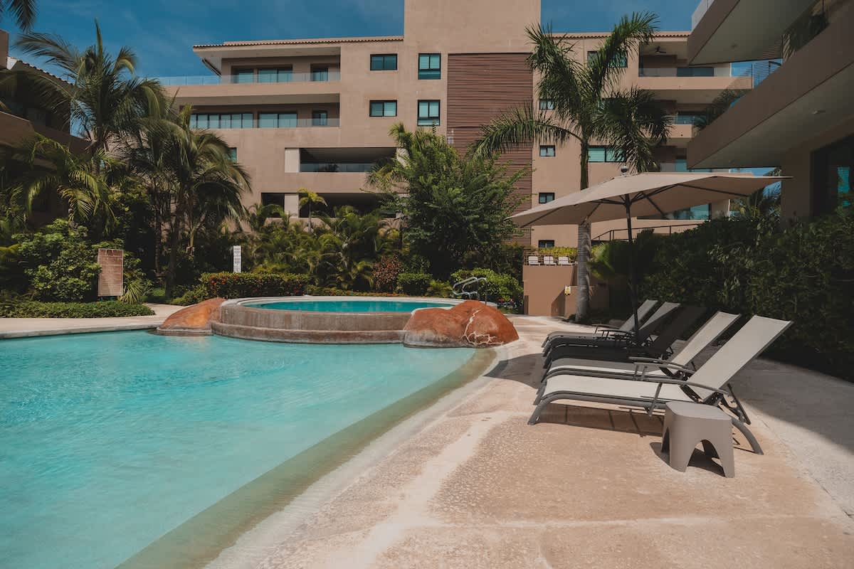 A serene swimming pool and circular hot tub with natural rock accents, flanked by a row of modern lounge chairs and a large patio umbrella, all set against a backdrop of lush tropical plants and a multi-story building.