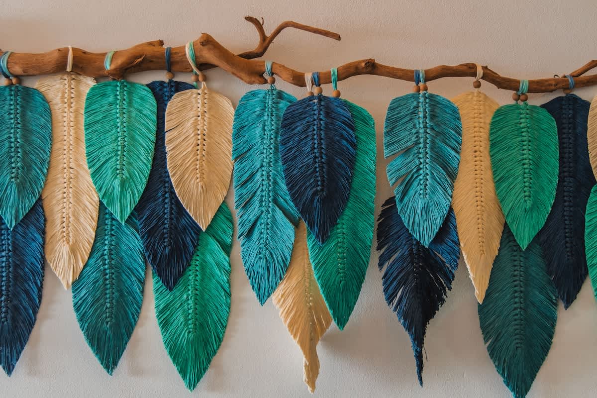 A vibrant macrame wall hanging featuring numerous feather-shaped elements in shades of teal, green, and beige, suspended from a natural wooden branch, adding a bohemian touch to the interior decor.