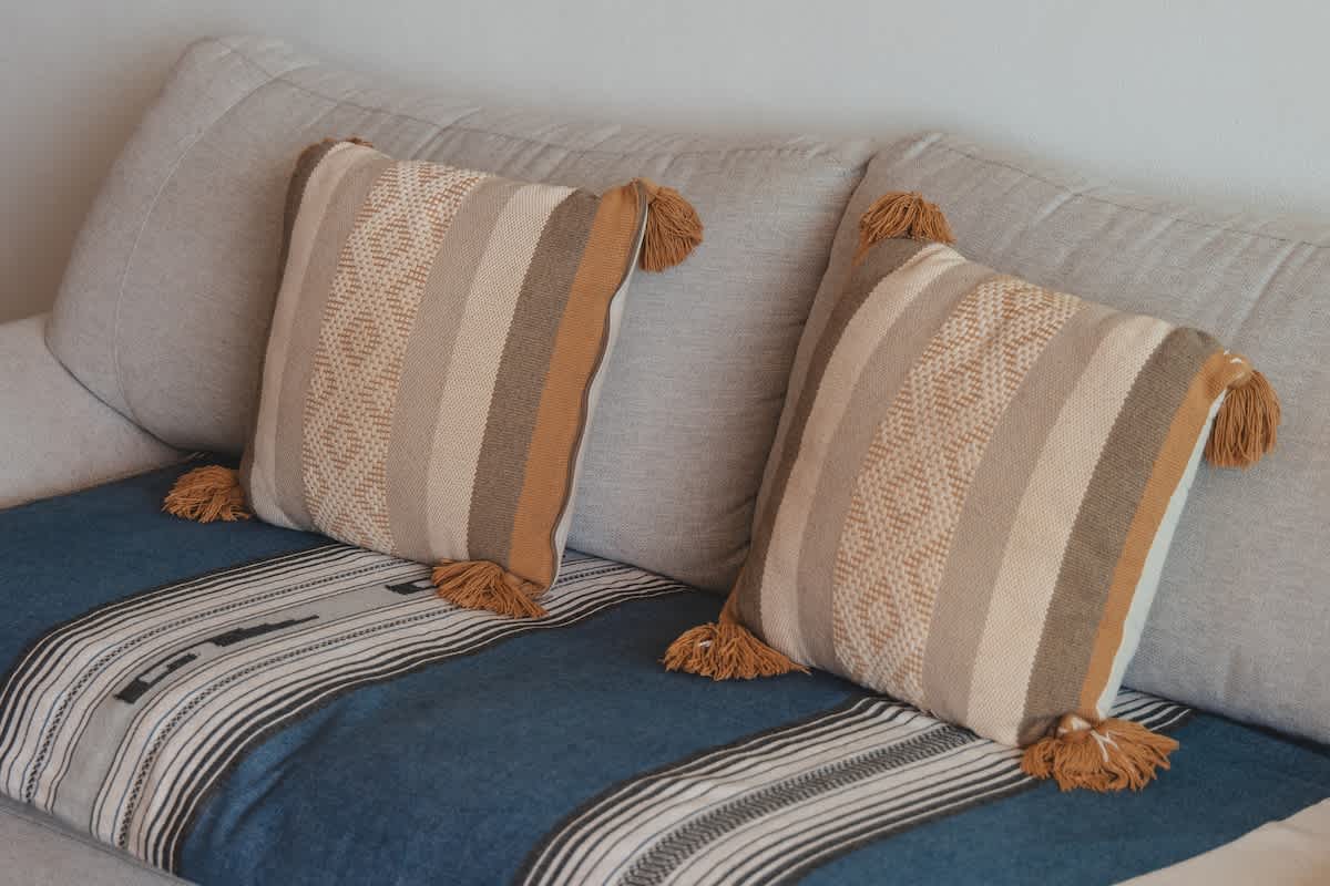 A pair of textured, striped throw pillows with decorative tassels in warm beige and brown tones are arranged on a light-colored sofa, accented by a blue and white patterned blanket.