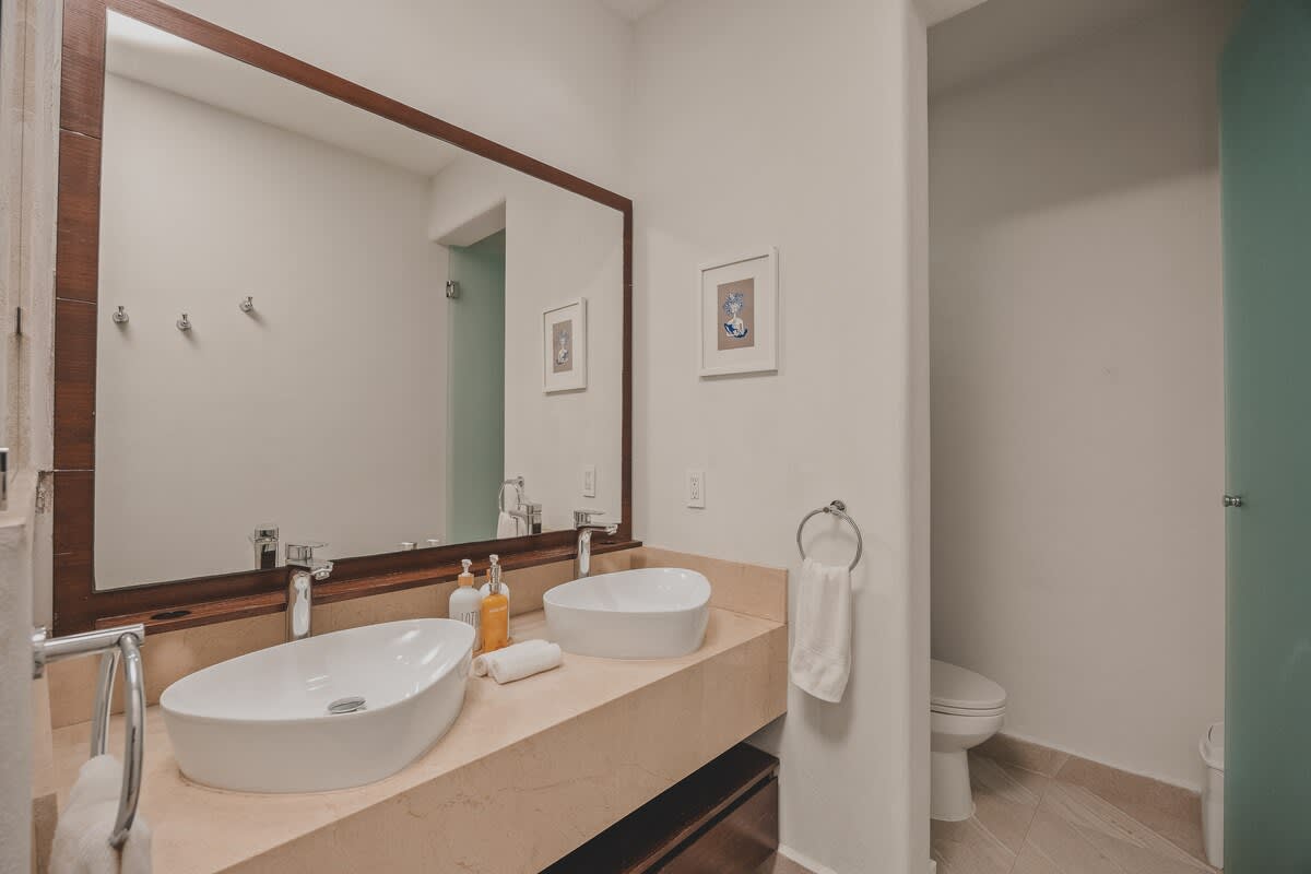 A clean and bright bathroom with a double vanity, vessel sinks, and a large mirror. The toilet is discreetly located in a separate alcove, accessible through a frosted glass door.
