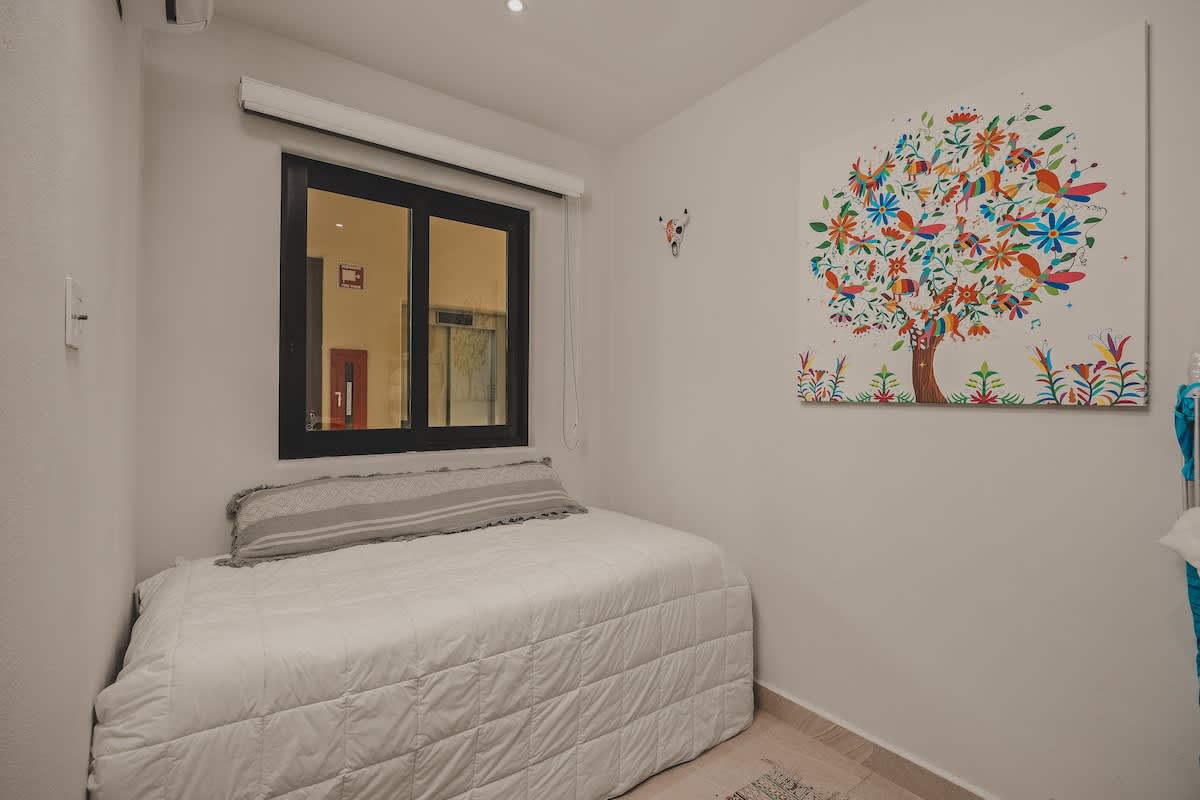 A compact bedroom featuring a single bed with white quilted bedding and a grey pillow, illuminated by a window with room-darkening shades. A vibrant, colorful tree artwork hangs on the wall, adding a cheerful touch.