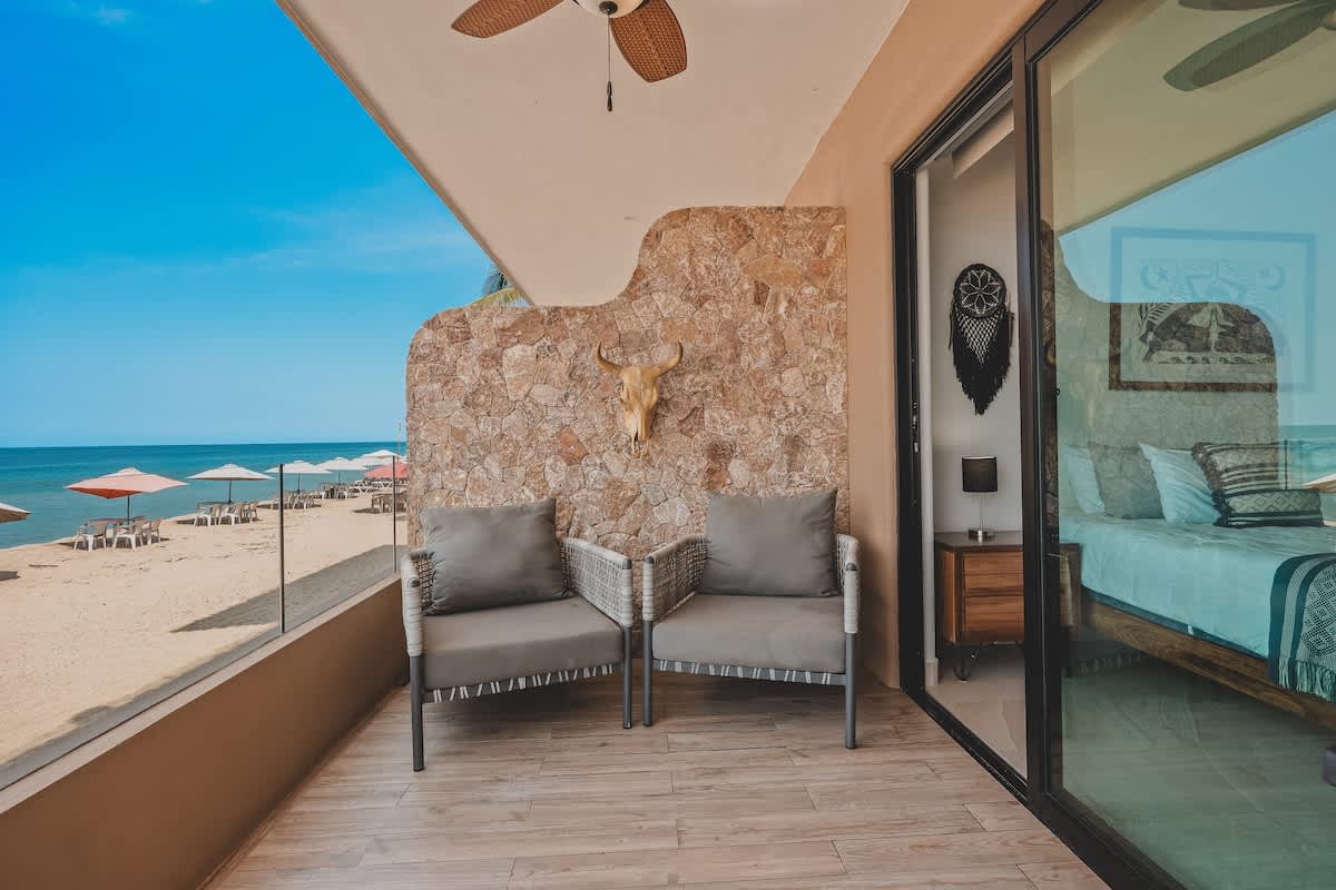 Balcony with two grey cushioned wicker chairs and a stone accent wall, offering a direct view of the sandy beach and ocean, with a glimpse of the bedroom interior through sliding glass doors.
