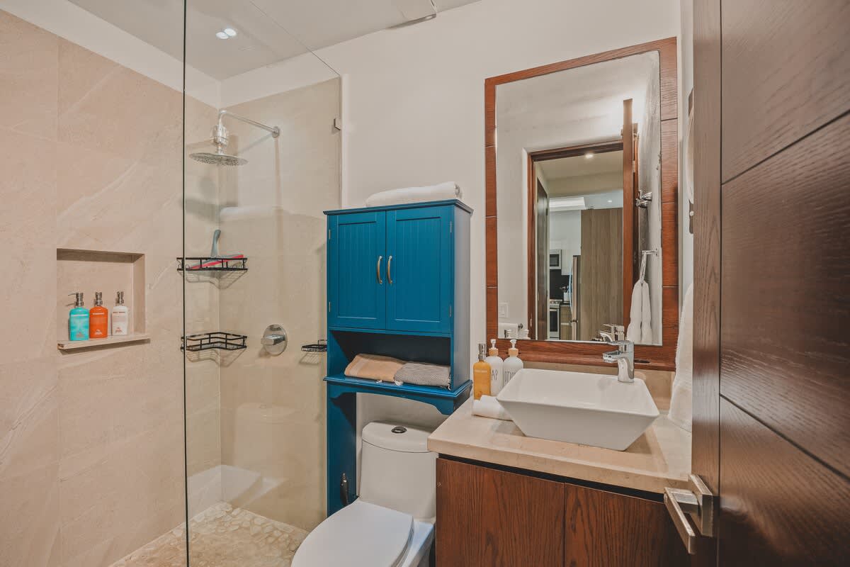 Modern bathroom with a walk-in shower featuring a rain showerhead and glass enclosure. It includes a white vessel sink on a vanity with a wooden mirror, and a vibrant blue storage cabinet above the toilet.