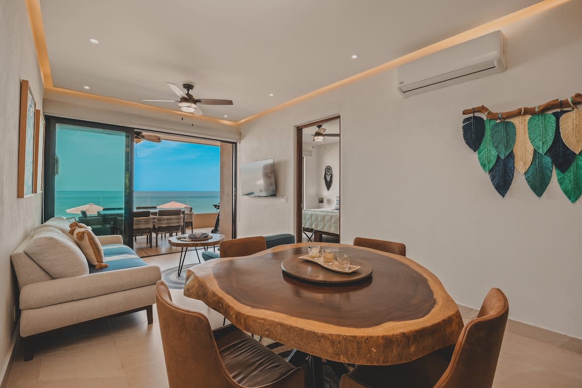 Open concept living and dining area with a large live-edge wood dining table, light-colored sofa, and a balcony overlooking the ocean. The room features recessed lighting, a ceiling fan, and a wall-mounted TV.