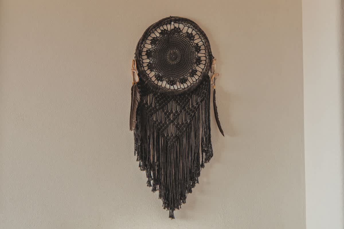 A dark, intricate macrame dreamcatcher with a circular woven center and long fringed tassels hangs against a light-colored wall, featuring some feather accents.
