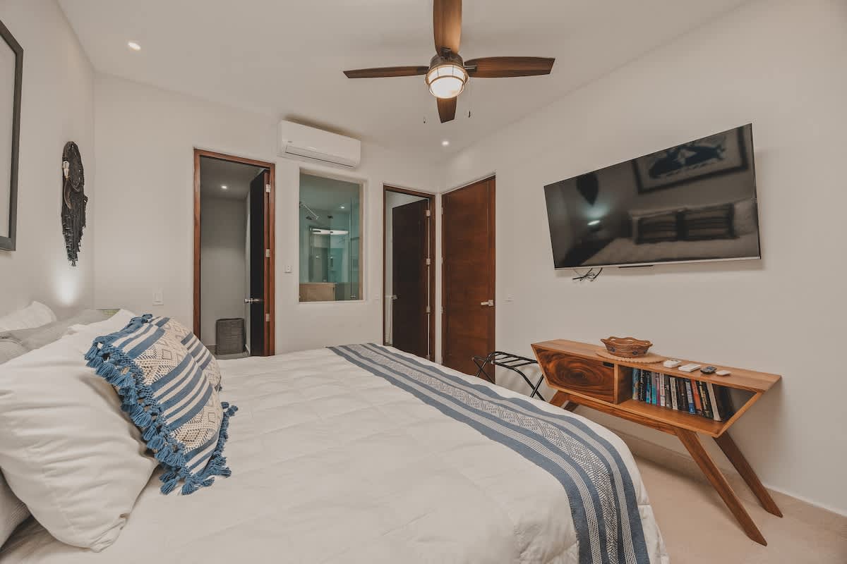 Another view of the master bedroom, showcasing a wall-mounted flat-screen TV above a wooden console table filled with books. The king bed with blue and white bedding is partially visible, along with access to an ensuite bathroom.