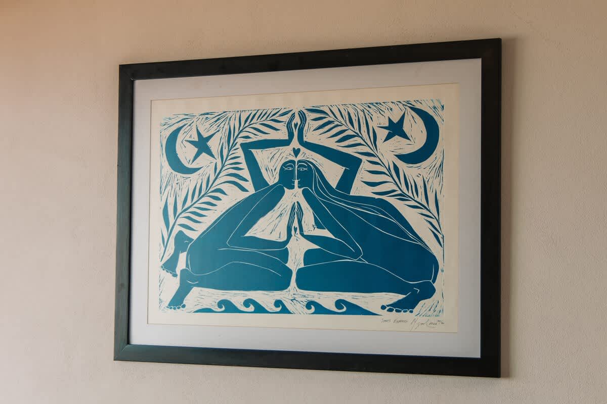 A framed piece of blue and white linocut art depicting two stylized figures in a meditative pose, surrounded by moon, stars, and waves, hanging on a light-colored wall, contributing to the room's artistic ambiance.