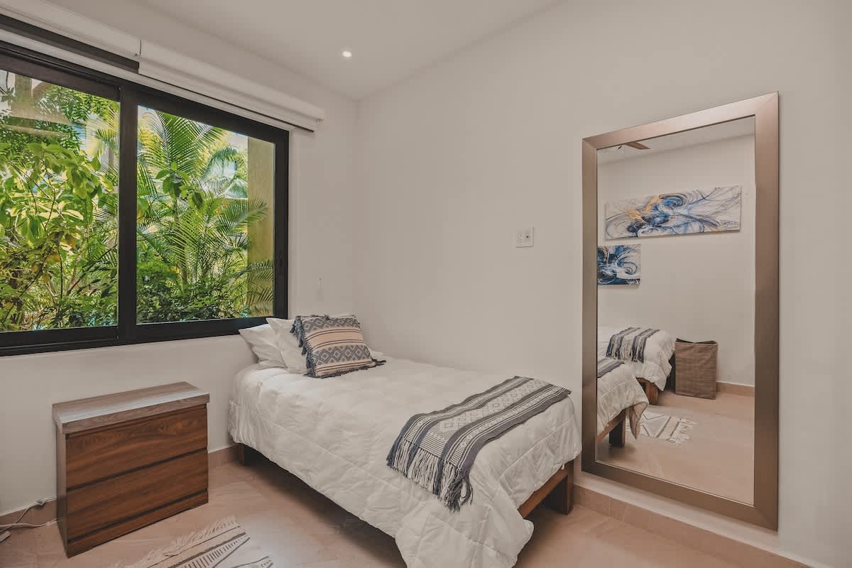 A bright bedroom corner with a single twin bed, white bedding, and a patterned throw, positioned next to a large window revealing vibrant green foliage. A tall, full-length mirror reflects the room's abstract artwork.