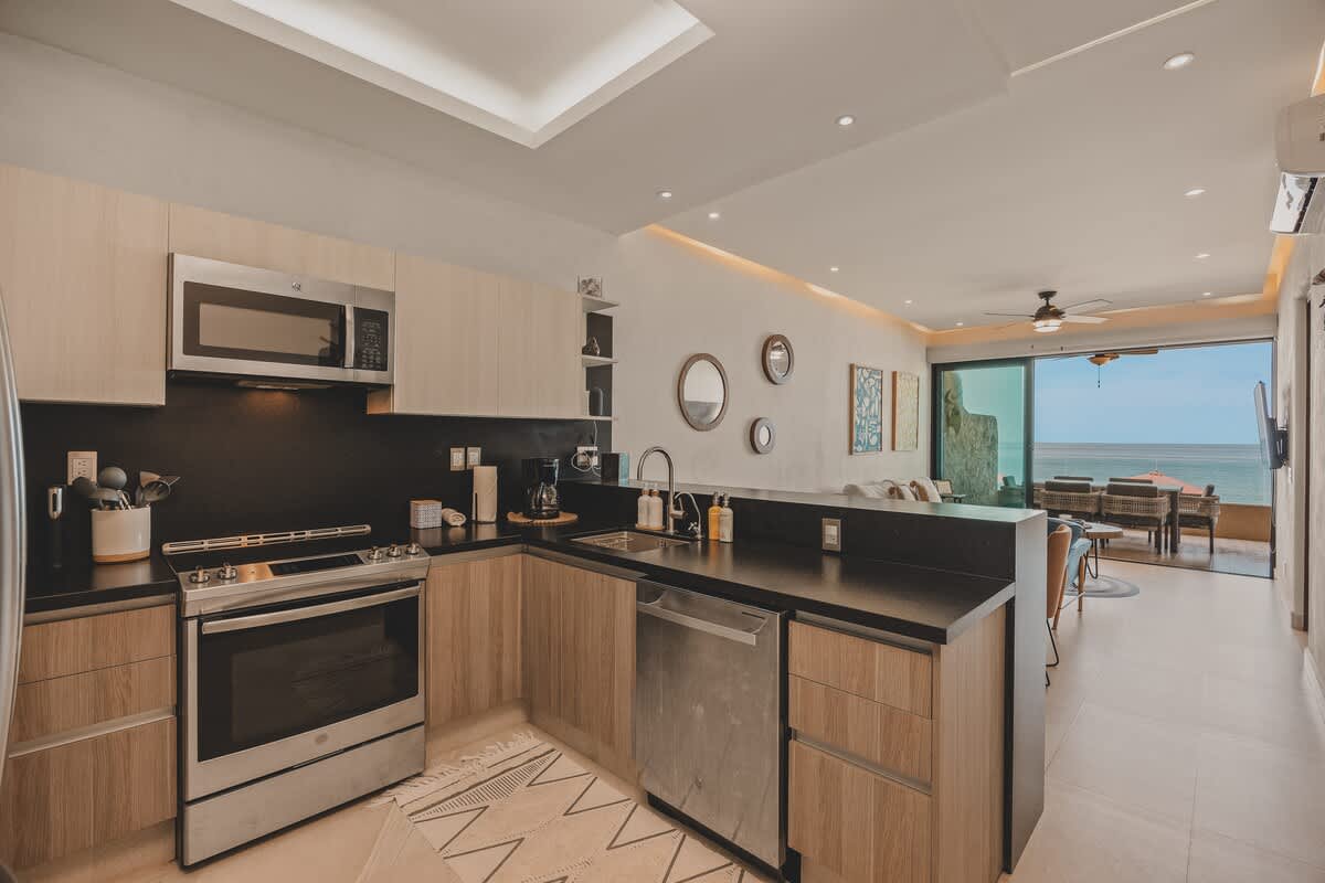 View of the kitchen from the dining area, showcasing the open layout, light wood cabinets, dark countertops, and stainless steel appliances. The living area and ocean view are visible in the background.
