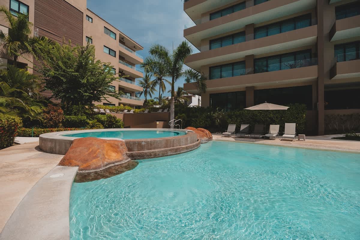 A resort-style swimming pool with a connected circular hot tub, surrounded by concrete decking, lush tropical landscaping, and modern multi-story buildings under a bright sky.