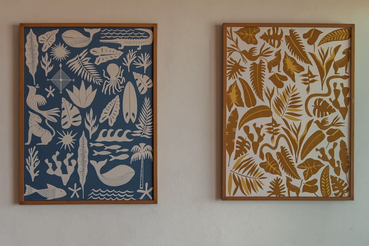 Two framed art prints with a tropical motif hang on a white wall; one features blue and white marine life and plants, the other gold and white jungle foliage and animals.