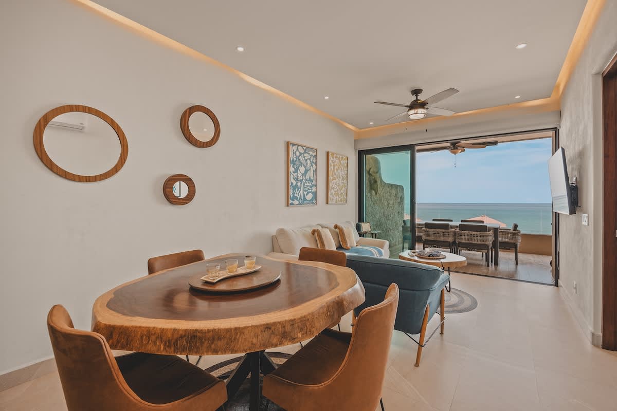 Another view of the open concept living and dining area, featuring a live-edge wood dining table, a light-colored sofa, and a balcony with ocean views. The wall is decorated with round mirrors and framed abstract art.