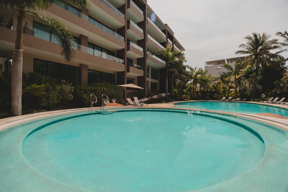 An expansive outdoor swimming pool area featuring a large, irregularly shaped pool with multiple entry points, surrounded by lush tropical plants and modern apartment buildings. Several lounge chairs are visible for relaxation.