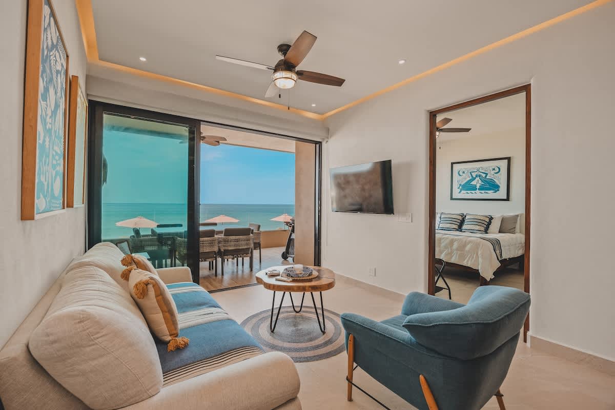 The living room opens to a balcony with stunning ocean views, featuring a light-colored sofa and a blue armchair. A wall-mounted TV is visible, and the space is bright with natural light.