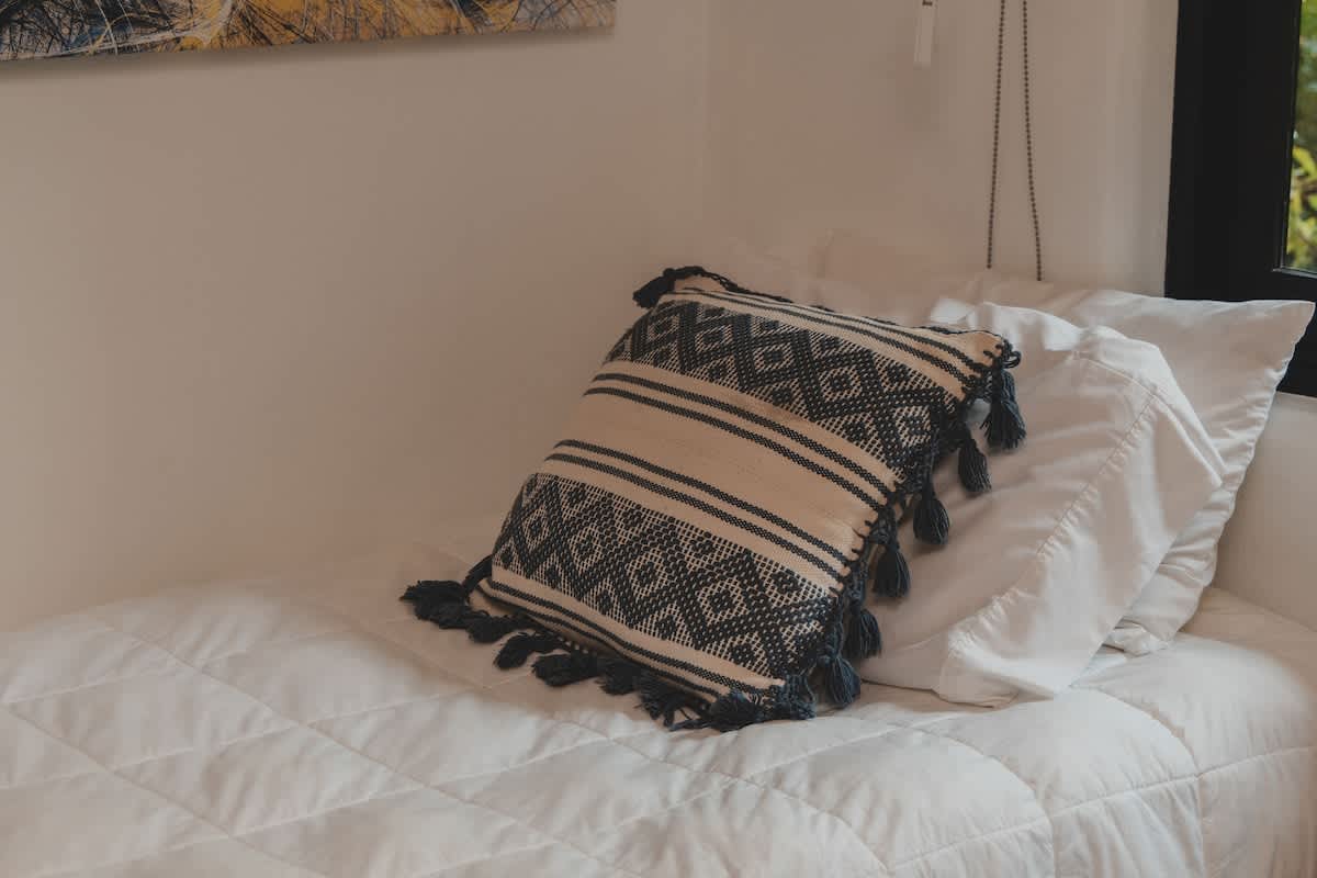 A cozy bed with crisp white quilted bedding, accented by a decorative throw pillow featuring a black and white geometric pattern and playful tassels. A window is partially visible in the background.