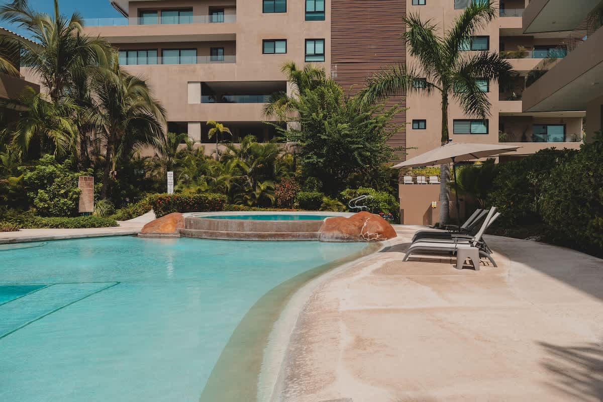 A curved swimming pool with a circular hot tub featuring rock accents, surrounded by light-colored concrete decking, several lounge chairs with an umbrella, and dense tropical greenery.