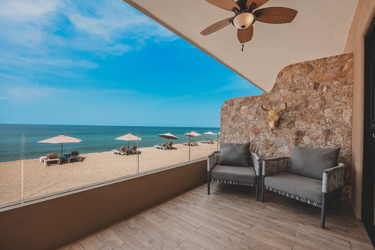 Balcony seating area with two grey cushioned wicker chairs, a stone accent wall with a bull skull, and a ceiling fan, offering expansive views of the sandy beach and blue ocean.