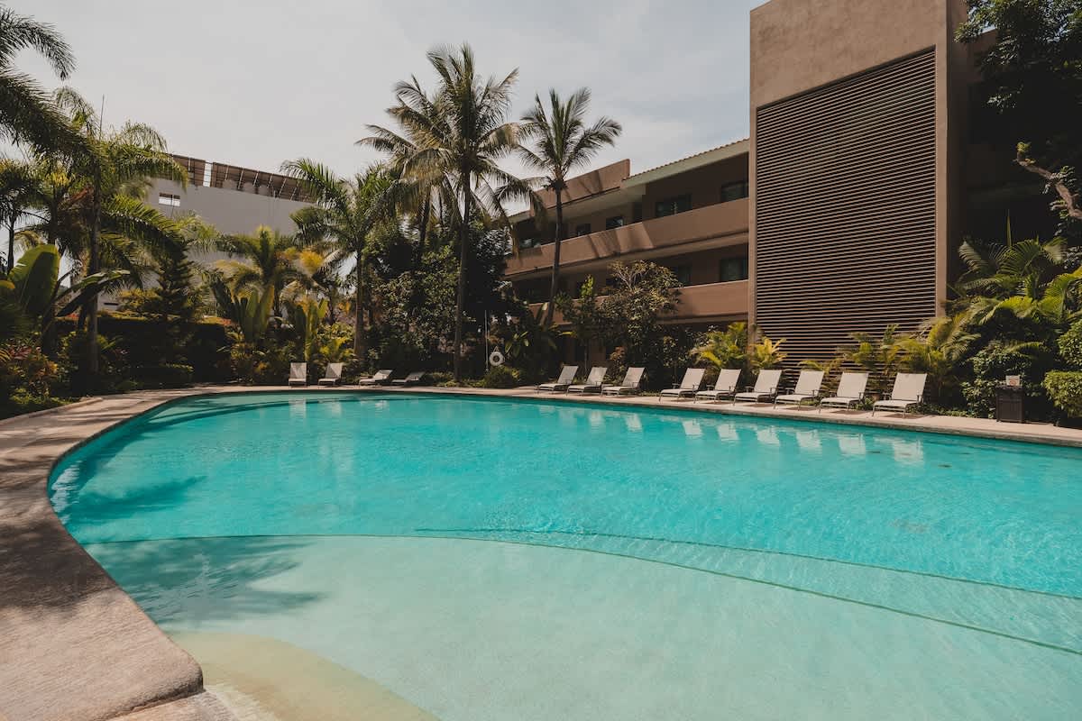Expansive swimming pool area featuring a curved pool, numerous lounge chairs lined along the edge, and tropical palm trees, set against the backdrop of the property's buildings.