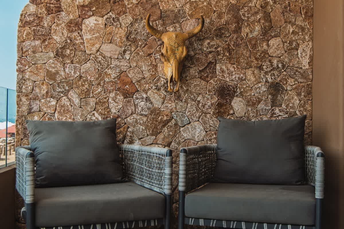 An inviting outdoor seating area featuring two modern grey armchairs with dark cushions, set against a rustic stone wall adorned with a decorative bull skull. A glimpse of the ocean is visible to the left, suggesting a balcony setting.