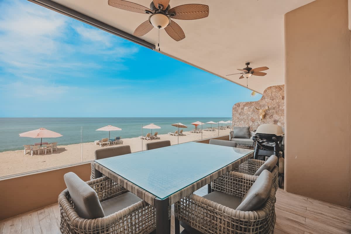 Spacious balcony with an outdoor dining table and chairs, a barbecue grill, and a stone accent wall, providing an unobstructed panoramic view of the sandy beach and the vast blue ocean.