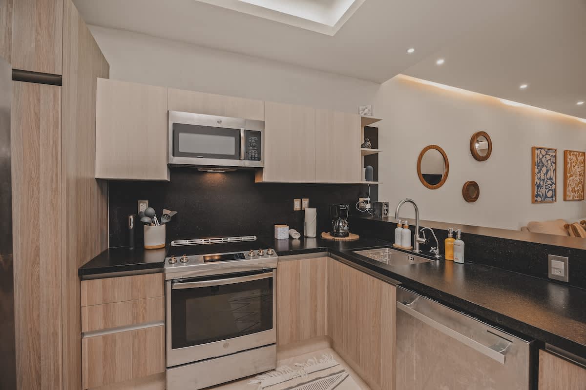 Another angle of the modern kitchen, showing the stainless steel oven and microwave, dark backsplash, light wood cabinets, and a sink with a dishwasher. A coffee maker and other kitchen items are on the counter.