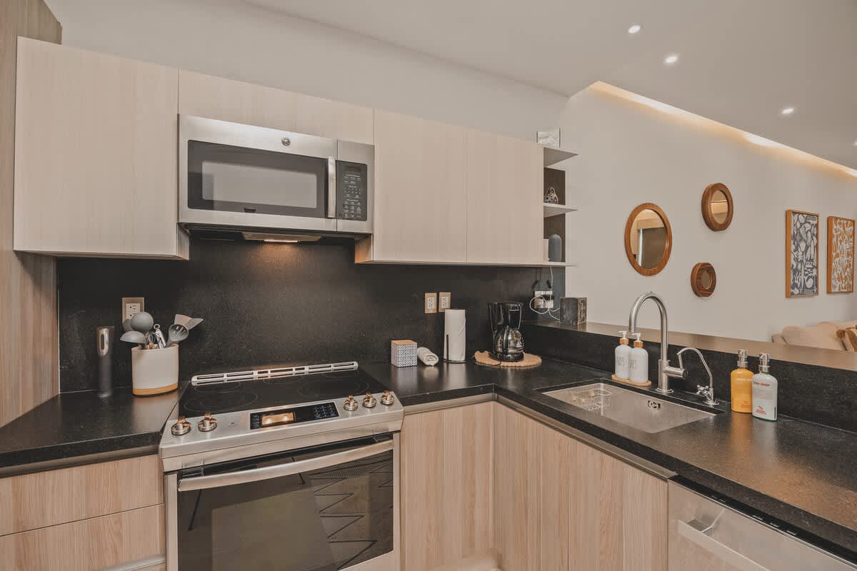 Detailed view of the kitchen counter, showing the electric range, built-in microwave, dark backsplash, and various kitchen essentials like a coffee maker, utensils, and cleaning supplies. The sink is also visible.