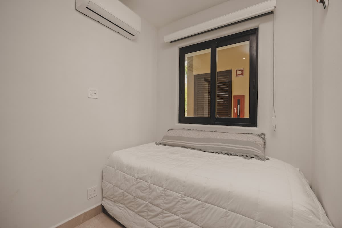 Close-up view of a single bed with white quilted bedding and a grey pillow in a bright bedroom, featuring a window with room-darkening shades. An air conditioning unit is mounted above the window.