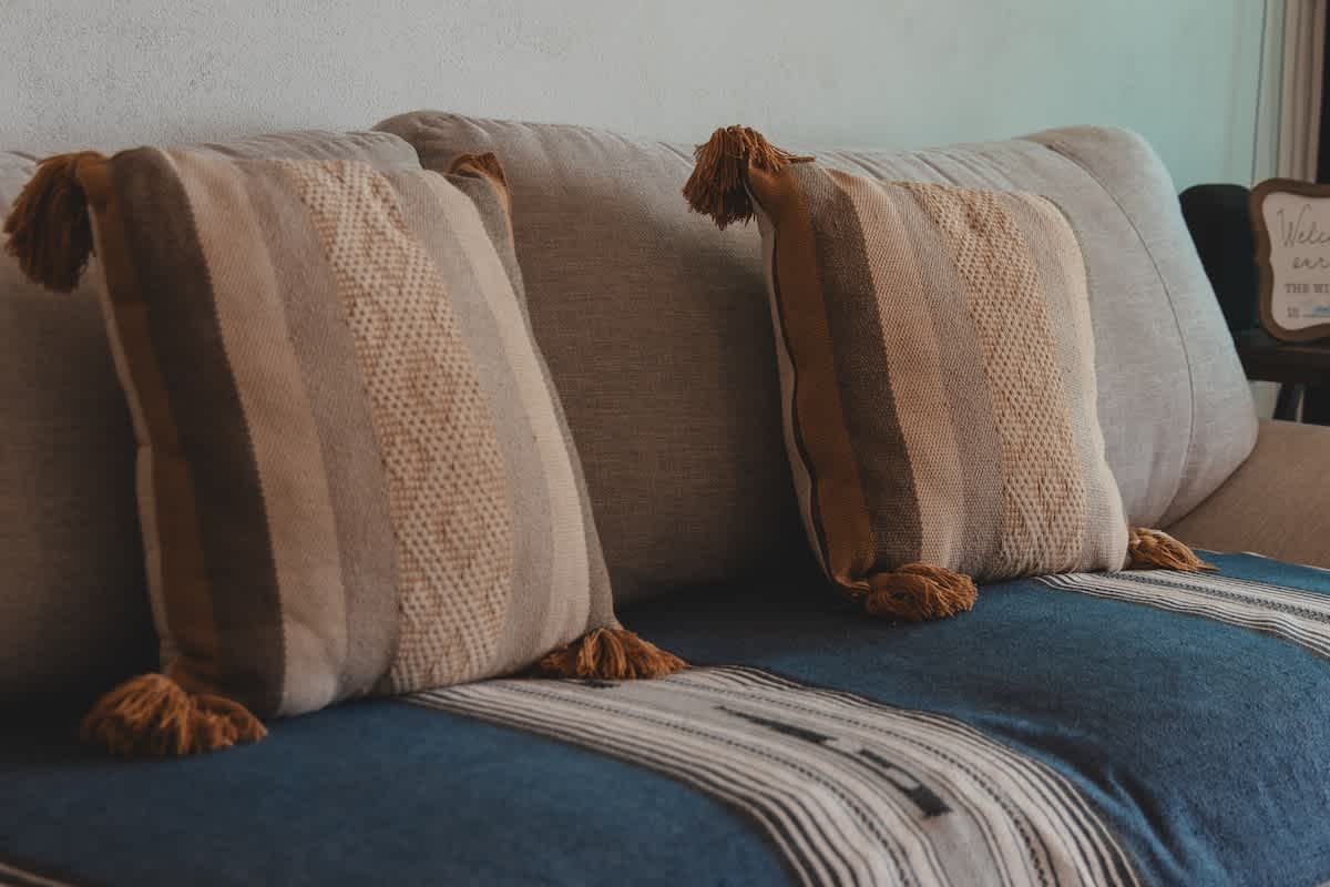 Two striped decorative pillows with tassels in earthy tones rest on a light grey sofa, which is covered with a blue and white patterned throw blanket, creating a cozy and inviting seating area.