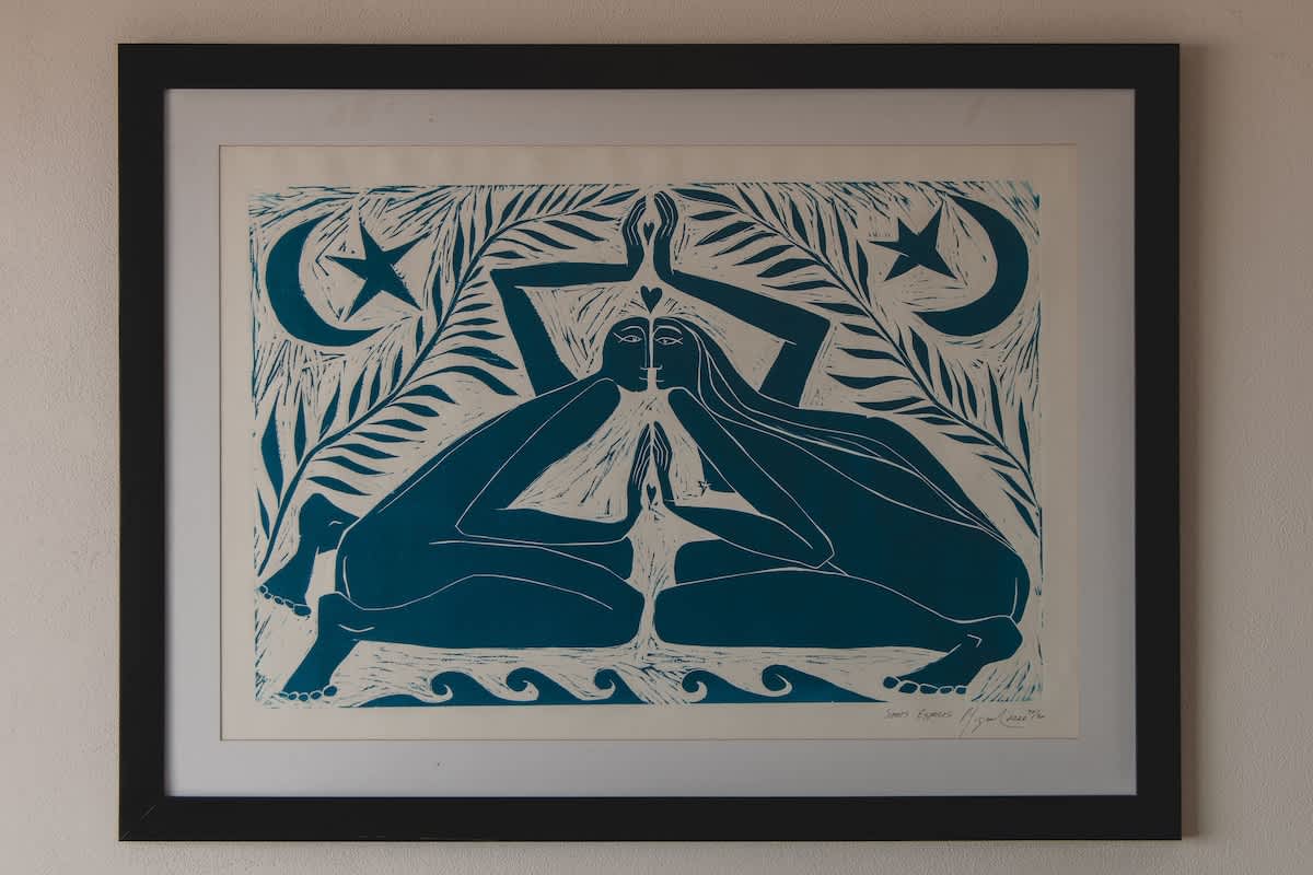 A framed piece of art depicts two stylized figures in blue and white, sitting opposite each other with hands raised, surrounded by tropical leaves, moons, and stars, set against a light wall.