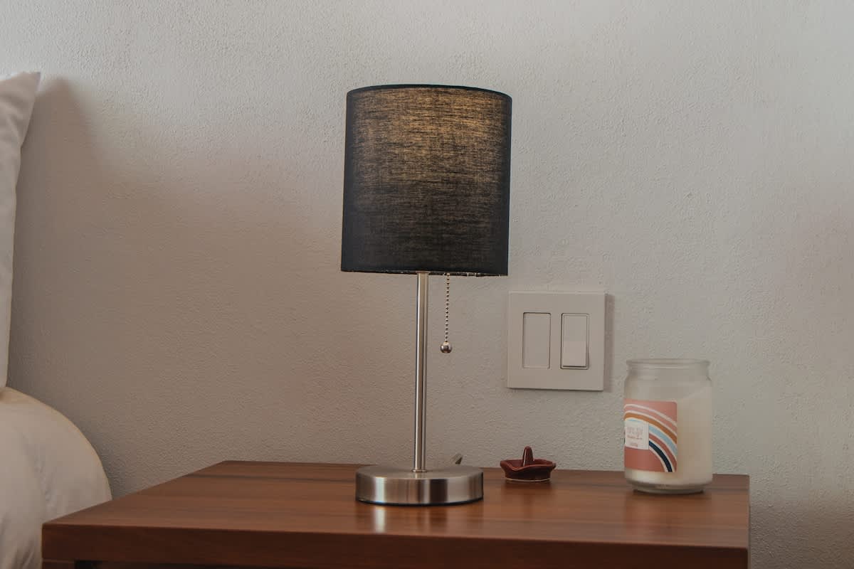 A modern silver-based table lamp with a dark fabric shade sits on a wooden nightstand next to a white wall with light switches, accompanied by a glass jar candle and a small decorative dish.