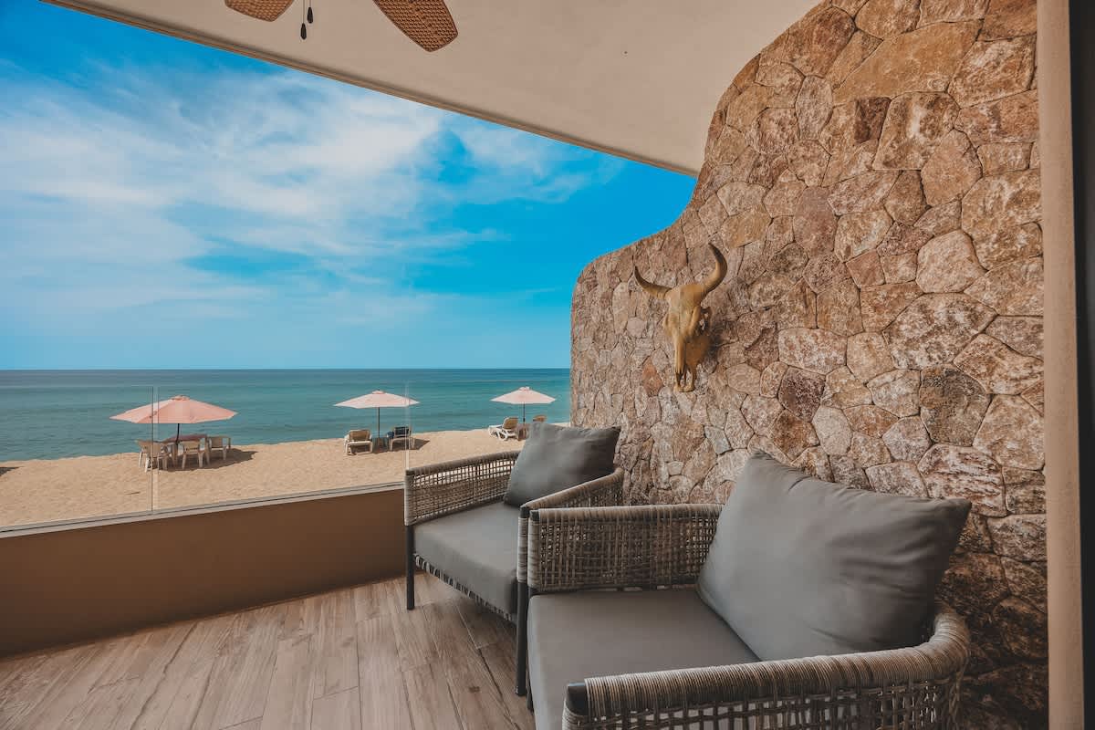 Balcony seating area featuring two grey cushioned wicker chairs, a prominent stone accent wall adorned with a bull skull, and a clear glass railing overlooking the beach and ocean.