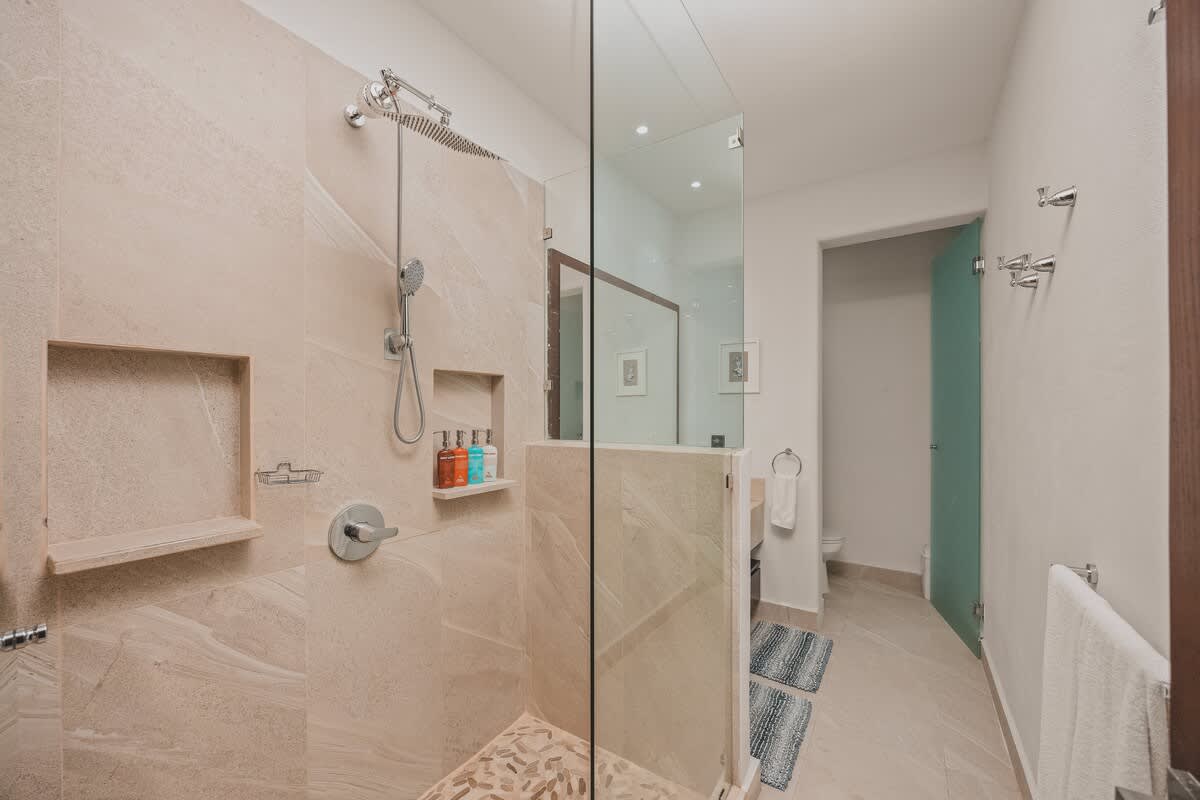 A close-up of the spacious walk-in shower, featuring light-colored tiled walls, a rain showerhead, and a built-in niche for toiletries. The floor has decorative pebble accents.