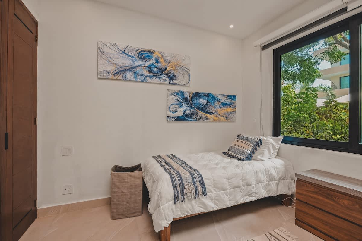 A cozy corner of a bedroom featuring a single twin bed with white bedding and a patterned throw, alongside a large window offering a view of lush greenery. Abstract blue and yellow artwork adorns the white wall.